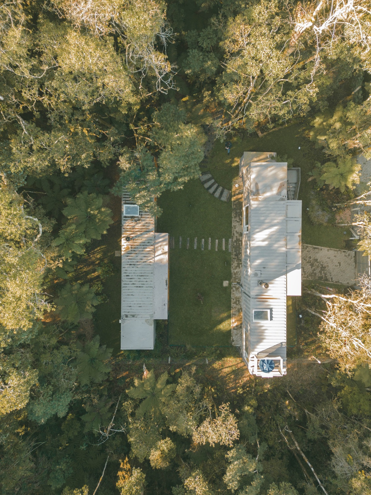 Vista aérea de cima dos dois containers da Kon na mata, ligados por um caminho de pedras no gramado, cercados por araucárias e samambaias